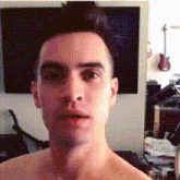 a man without a shirt is looking at the camera with a guitar hanging on the wall behind him