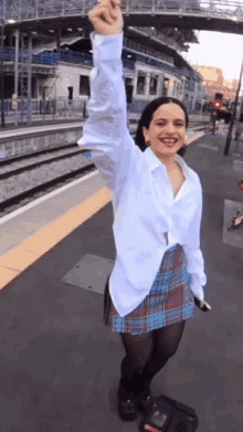 a woman wearing a plaid skirt and a white shirt stands on a train platform with her fist in the air