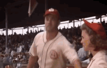 a man in a baseball uniform is standing next to a woman in a red hat in a baseball stadium .