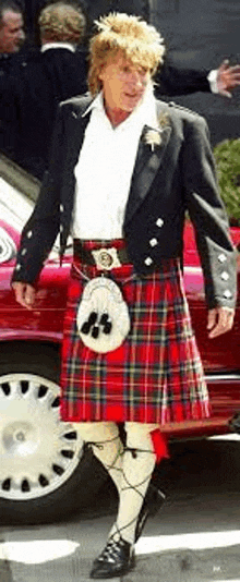 a man wearing a kilt and a black jacket is standing in front of a red car .