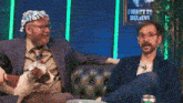 two men sitting on a couch with a sign that says i want to believe in the background