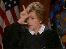 a woman in a judge 's robe is sitting in front of an american flag and making a funny face .