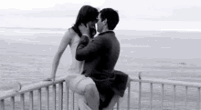 a man is sitting on a woman 's lap on a railing overlooking the ocean .