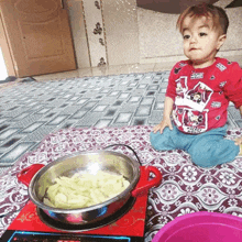 a child wearing a mickey mouse shirt sits on the floor next to a pot of food