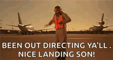 a man in an orange vest is standing on a runway with planes in the background .
