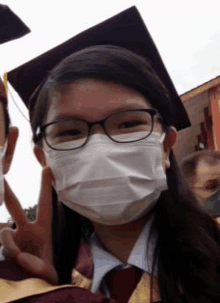 a girl wearing a graduation cap and gown is wearing a mask and giving a peace sign .