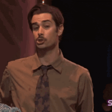 a man in a brown shirt and tie with the letter s on the bottom right