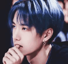 a young man with blue hair and earrings is sitting down with his hand on his chin .