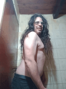 a shirtless man with long hair and glasses smiles in a bathroom