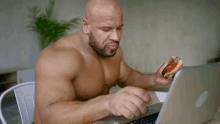 a shirtless man is eating a hot dog while looking at a laptop computer .