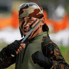 a man with camouflage paint on his face is holding a knife in his hand .