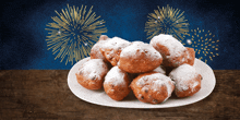 a plate of doughnuts with powdered sugar on a table with fireworks in the background