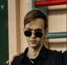 a young man wearing sunglasses and a black shirt is standing in front of a wooden wall .