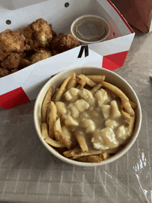 a bowl of french fries and gravy next to a box of fried chicken