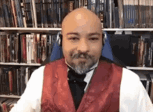 a bald man with a beard is wearing a red vest and bow tie while sitting in front of a bookshelf .