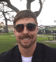 a man wearing sunglasses and a black jacket smiles in front of a picnic table