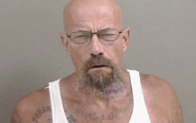 a bald man with a beard and glasses is wearing a white tank top and has tattoos on his chest .