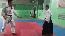 a man in a karate uniform is standing next to another man in a karate uniform on a mat in a gym .