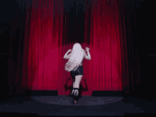 a woman with long white hair stands on a stage in front of a red curtain