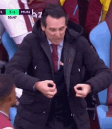 a man in a suit and tie is sitting in a stadium watching a soccer game with the score 3-1 mun