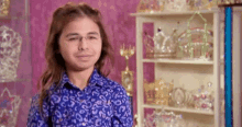 a young boy wearing glasses and a blue shirt is standing in front of a shelf full of tiaras .