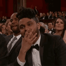 a man in a tuxedo and bow tie is covering his mouth with his hand while sitting in a crowd .