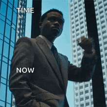 a man in a suit and tie stands in front of a window with the words time now above him