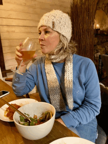 a woman drinking a glass of wine while sitting at a table with a bowl of food