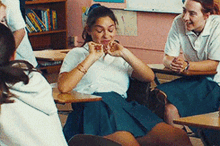 a group of girls are sitting at desks in a classroom and one girl is making a heart shape with her hands