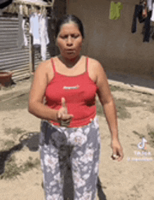 a woman in a red tank top and floral pants is giving a thumbs up in a yard .