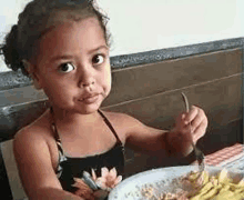 a little girl is sitting at a table with a plate of food and a fork in her hand .