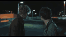 two men are standing next to each other in a parking lot at night and talking to each other .