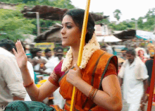 a woman in an orange saree is holding a yellow stick in front of a crowd and the name vonkat is on the bottom right