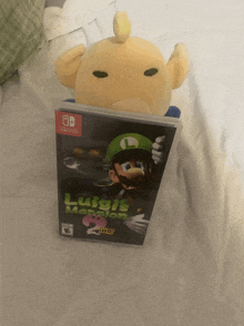 a luigi 's mansion 2 video game sits next to a stuffed animal on a bed