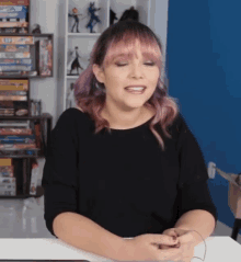 a woman with pink hair is sitting at a table in front of a shelf with a lot of books on it