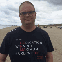 a man stands on a beach wearing a shirt that says all for dedication winning maximum hard work