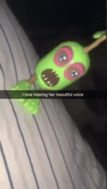 a green toy with red eyes is sitting on a bed with a caption that says i love hearing her beautiful voice