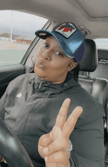 a woman in a black jacket and a blue hat is giving a peace sign in a car