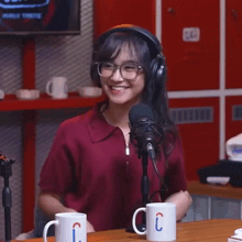 a woman wearing headphones and glasses smiles in front of a microphone next to two mugs with the letter c on them