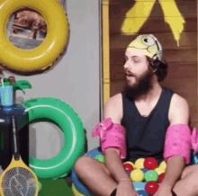 a man with a beard wearing a swim cap is sitting on a ball pit surrounded by inflatable balls .