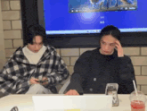 two young men are sitting at a table looking at their cell phones .