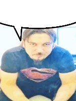 a man with a beard wearing a superman shirt is sitting down with a speech bubble above his head .