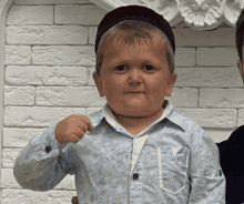 a small boy wearing a hat and a blue shirt is standing in front of a white brick wall .