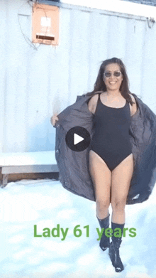 a woman in a black swimsuit and boots is standing in the snow with a play button .