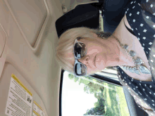 a woman wearing sunglasses and a necklace sits in a car with a warning sign on the visor