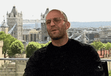 a man wearing sunglasses and a black shirt is sitting in front of a city with a bridge in the background .