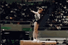 a female gymnast stands on a balance beam in front of a crowd with a green exit sign in the background