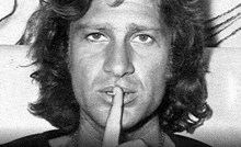 a man with long hair is holding his finger to his mouth and making a silence sign .