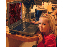 a little girl sits at a table with a laptop and a can of paint next to her