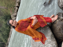 a woman in an orange and red dress sits on a rock by a river